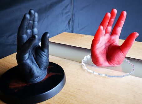 Zwei in Epoxidharz abgeformte Hände. Die Finger sind leicht abgespreizt. Dabei ist eine Rot und die zweite schwarz.