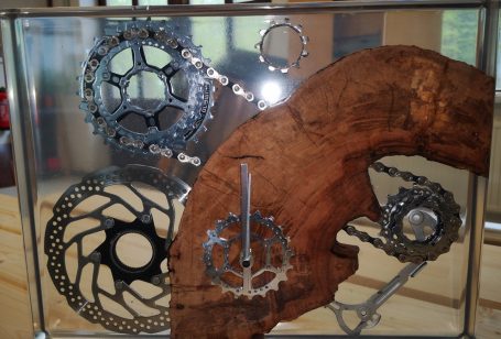 Deko Bike für Fahrradenthusiasten. Chromglänzende Fahrradteile wie Zahnräder und Kette in rötlichen Holz in klarem Epoxidharz