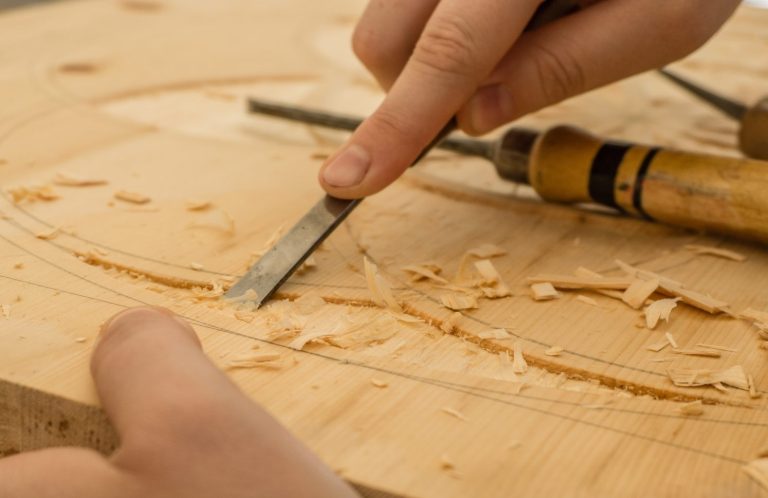 Eine Hand verziert Holz mit einem Meißel, Späne liegen verstreut auf der Oberfläche.